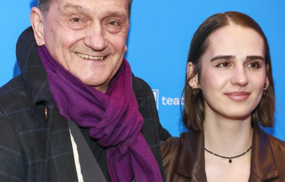 Starszy mężczyzna w ciemnym płaszczu i fioletowym szalu stoi obok młodej kobiety z krótkimi, prostymi włosami; oboje patrzą w kierunku kamery i uśmiechają się lekko na tle niebieskiej ściany z białymi napisami.