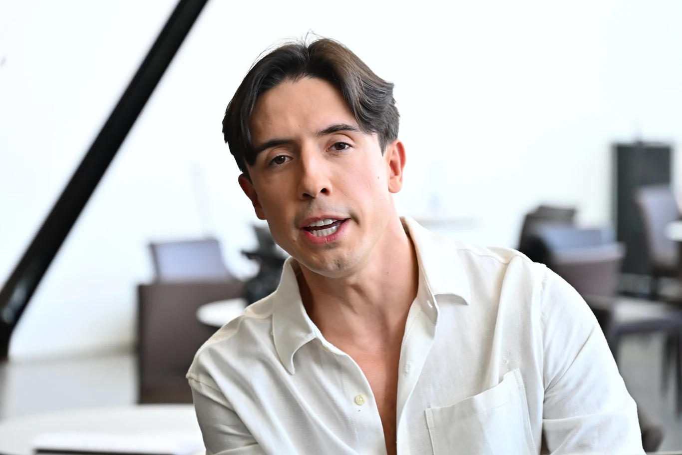 Dawid Kwiatkowski Dawid Kwiatkowski w białej koszuli siedzi w jasnym wnętrzu, w tle krzesła i stoliki, skupienie na twarzy