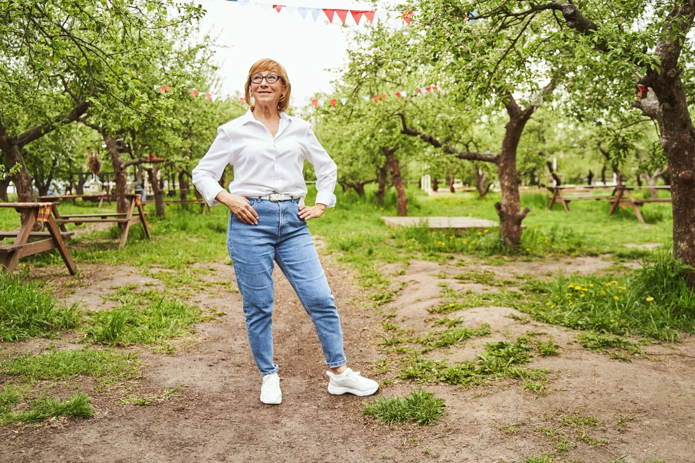 Zbyt swobodny strój także nie jest dobrym wyjściem kobieta w okularach, białej koszuli i dżinsach stoi pewnie na leśnej ścieżce wśród drzew owocowych, w tle widać drewniane stoły i ławki oraz wiszące girlandy z kolorowych trójkątnych flag
