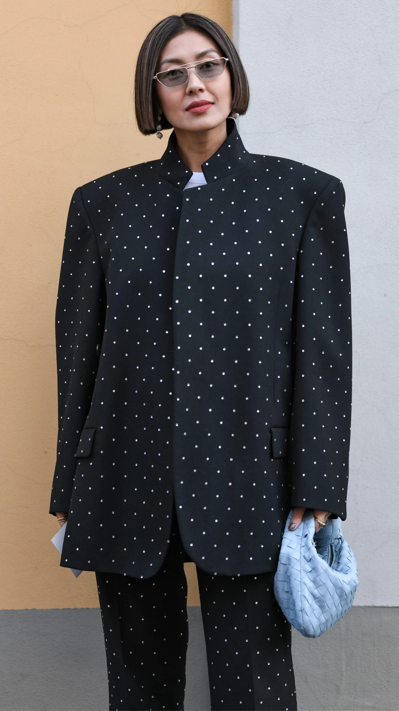 Kobieta w okularach przeciwsłonecznych ubrana w czarny, oversize'owy garnitur z drobnym, białym wzorem, trzymająca jasnoniebieską, pikowaną torebkę w lewej ręce, na tle dwukolorowej ściany.