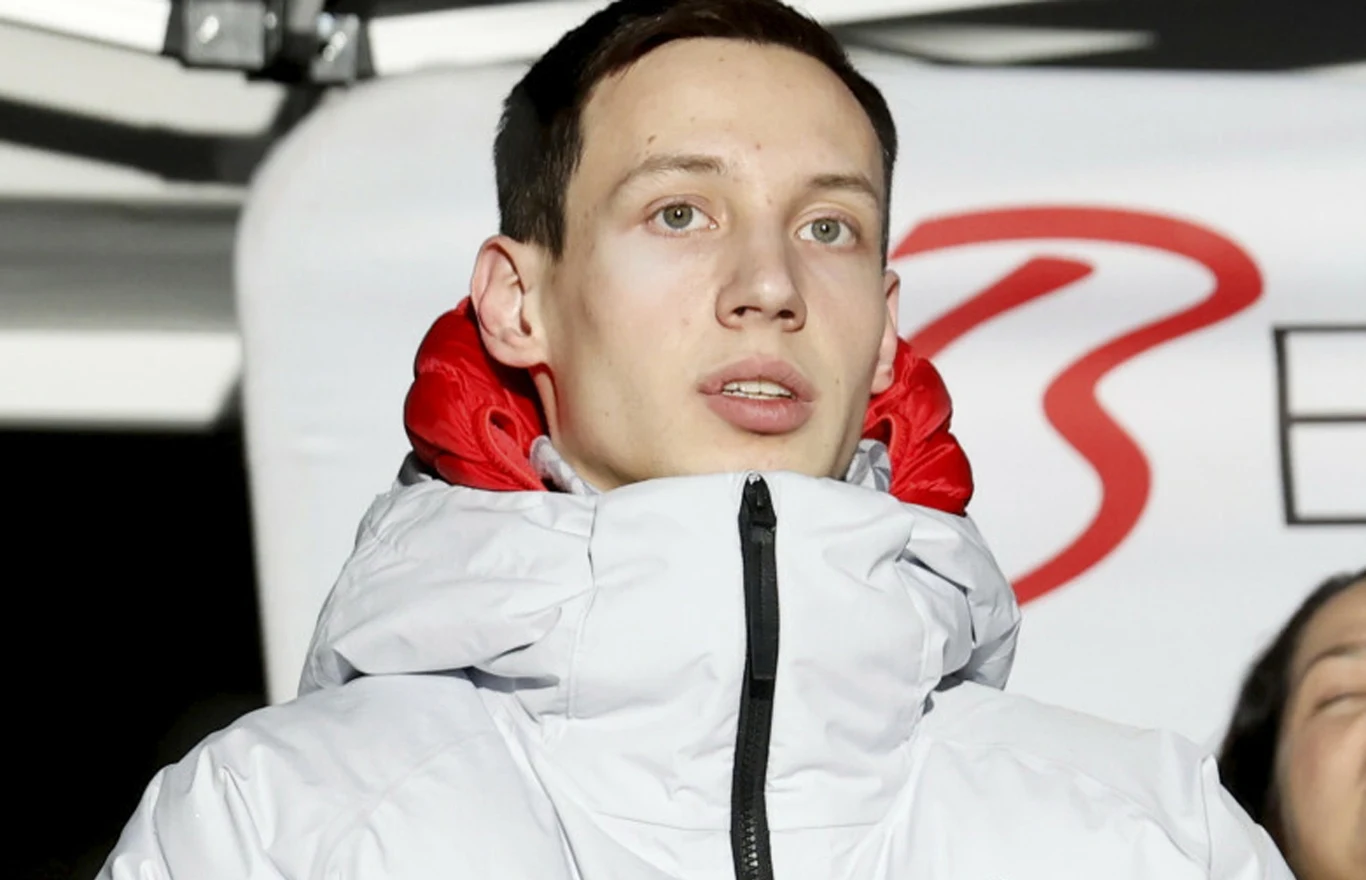Kacper Tomasiak po powrocie z zimowych igrzysk olimpijskich Mediolan-Cortina 2026 młody mężczyzna w białej kurtce z czerwonym podszyciem patrzący przed siebie, tło z elementem dużej litery 'B'