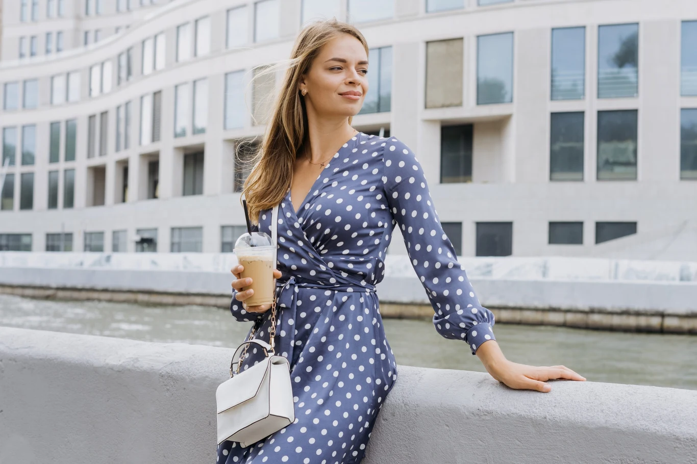kobieta w niebieskiej sukience w białe grochy, z białą torebką na ramieniu, oparta o balustradę nad kanałem, trzyma w dłoni kawę mrożoną, za nią nowoczesny budynek z dużymi oknami