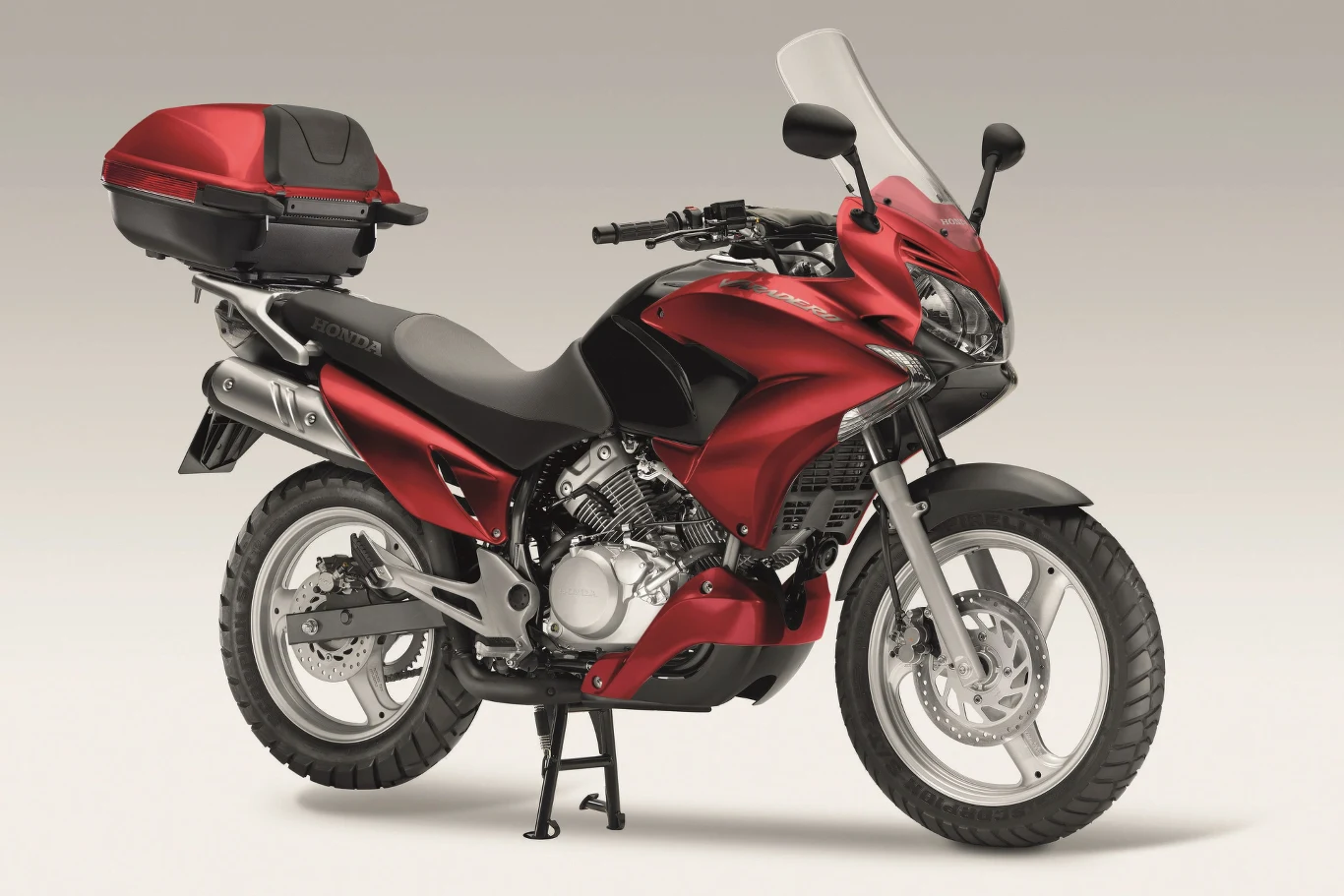 Honda XL 125V Varadero czerwony motocykl turystyczny z dużą szybą przednią, kufrem zamontowanym z tyłu i srebrnym wydechem, stojący na centralnej podstawce na jasnym tle