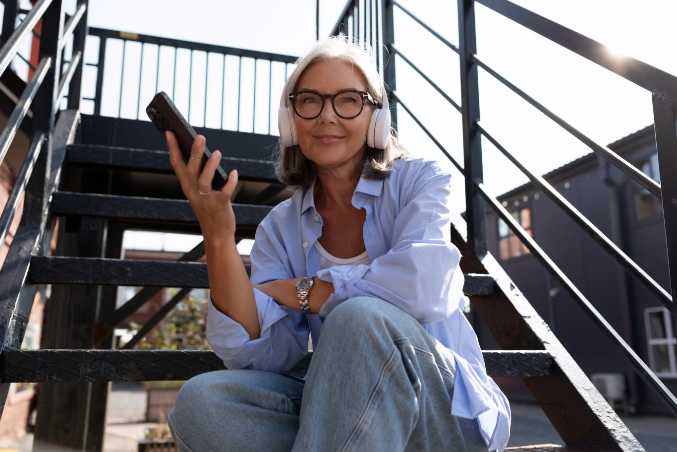 Uśmiechnięta kobieta z siwymi włosami i okularami siedzi na schodach zewnętrznych, trzyma w dłoni smartfon i ma na uszach słuchawki, ubrana w luźną koszulę i dżinsy.