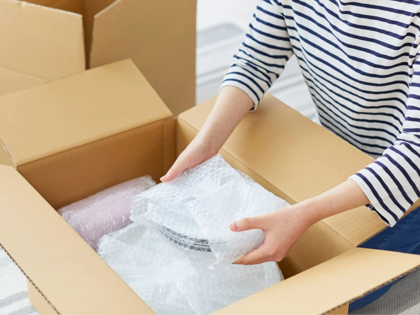 Jak przygotować się do przeprowadzki? Osoba pakująca talerze owinięte folią bąbelkową do kartonowego pudełka, przygotowując się do przeprowadzki