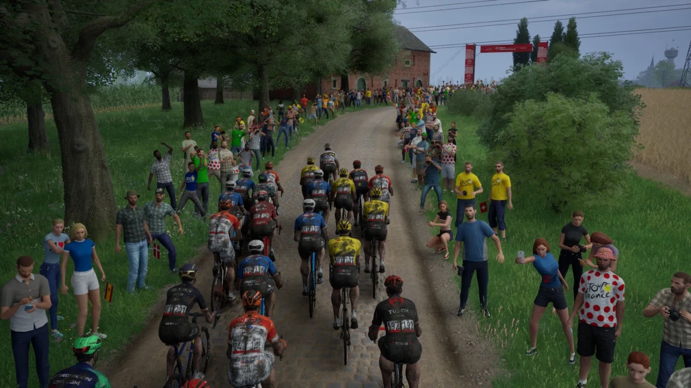 Tour de France 2026 peleton kolarzy jadących w wyścigu szosowym po wąskiej, brukowanej drodze otoczonej licznymi kibicami stojącymi po obu stronach trasy; w tle charakterystyczna wiejska zabudowa oraz baner oznaczający etap wyścigu