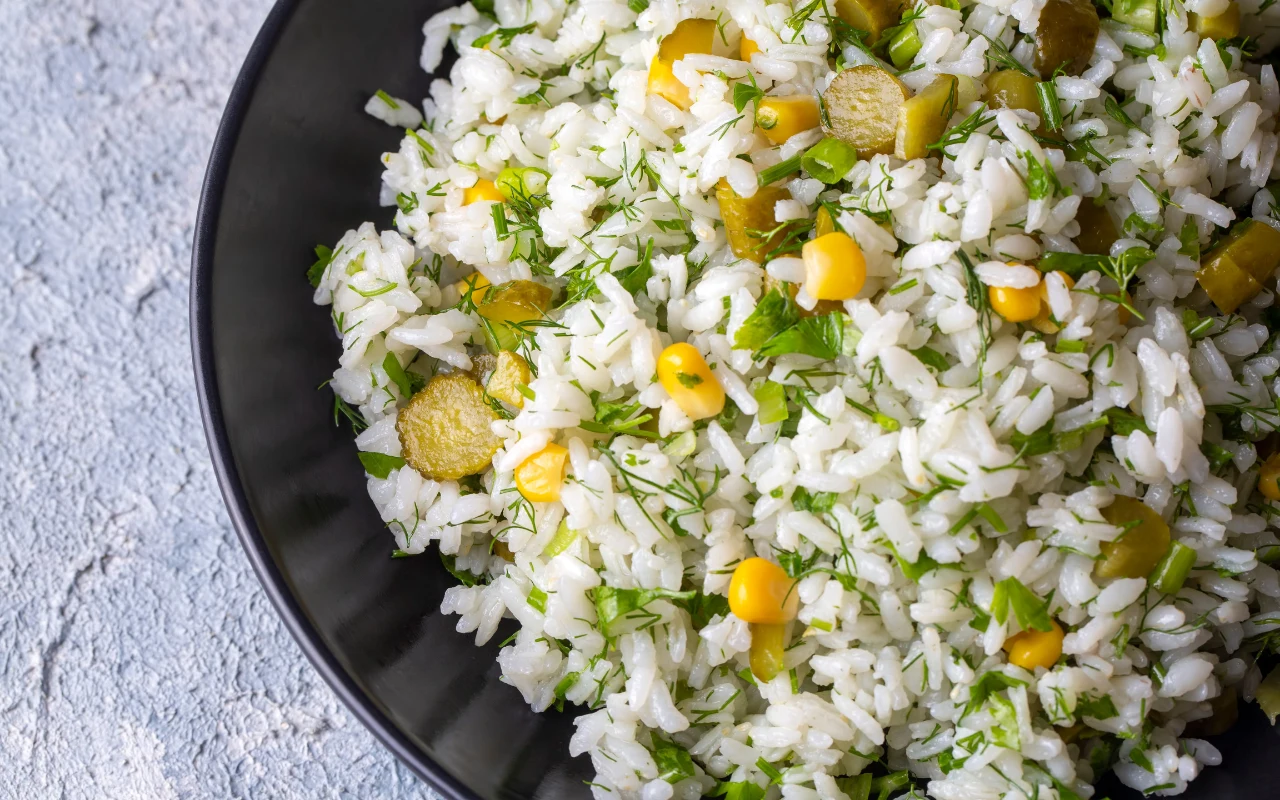 Sałatka na bazie ryży lub makaronu to świetny sposób na szybką kolację bez chleba sałatka z białego ryżu z dodatkiem zielonego koperku, kukurydzy i pokrojonych ogórków konserwowych, podana na czarnym talerzu