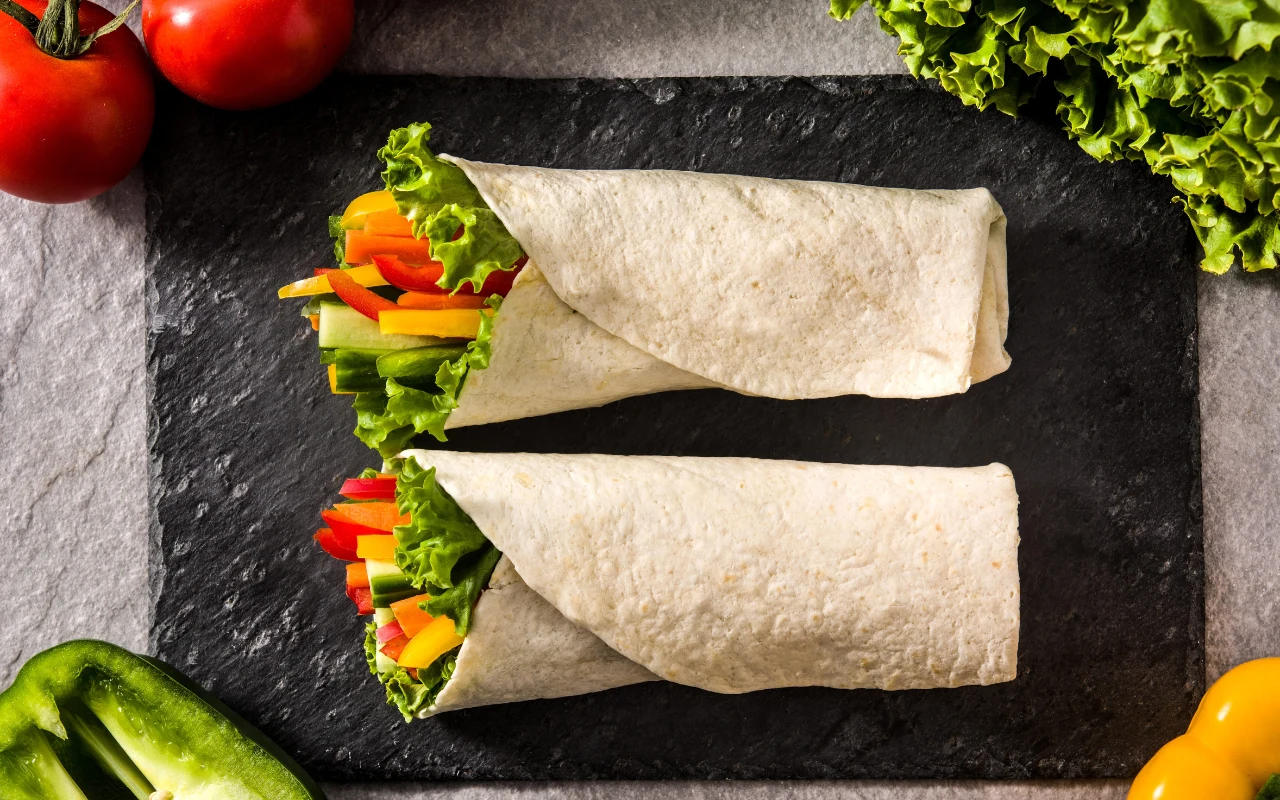 W tortillę zawinąć można prawie wszystko. To dobry pomysł na kolację bez chleba Dwie tortille zawinięte z sałatą i kolorowymi warzywami, takimi jak papryka oraz marchewka, ułożone na prostokątnej, czarnej desce; w tle widoczne kawałki sałaty, pomidory i papryka.