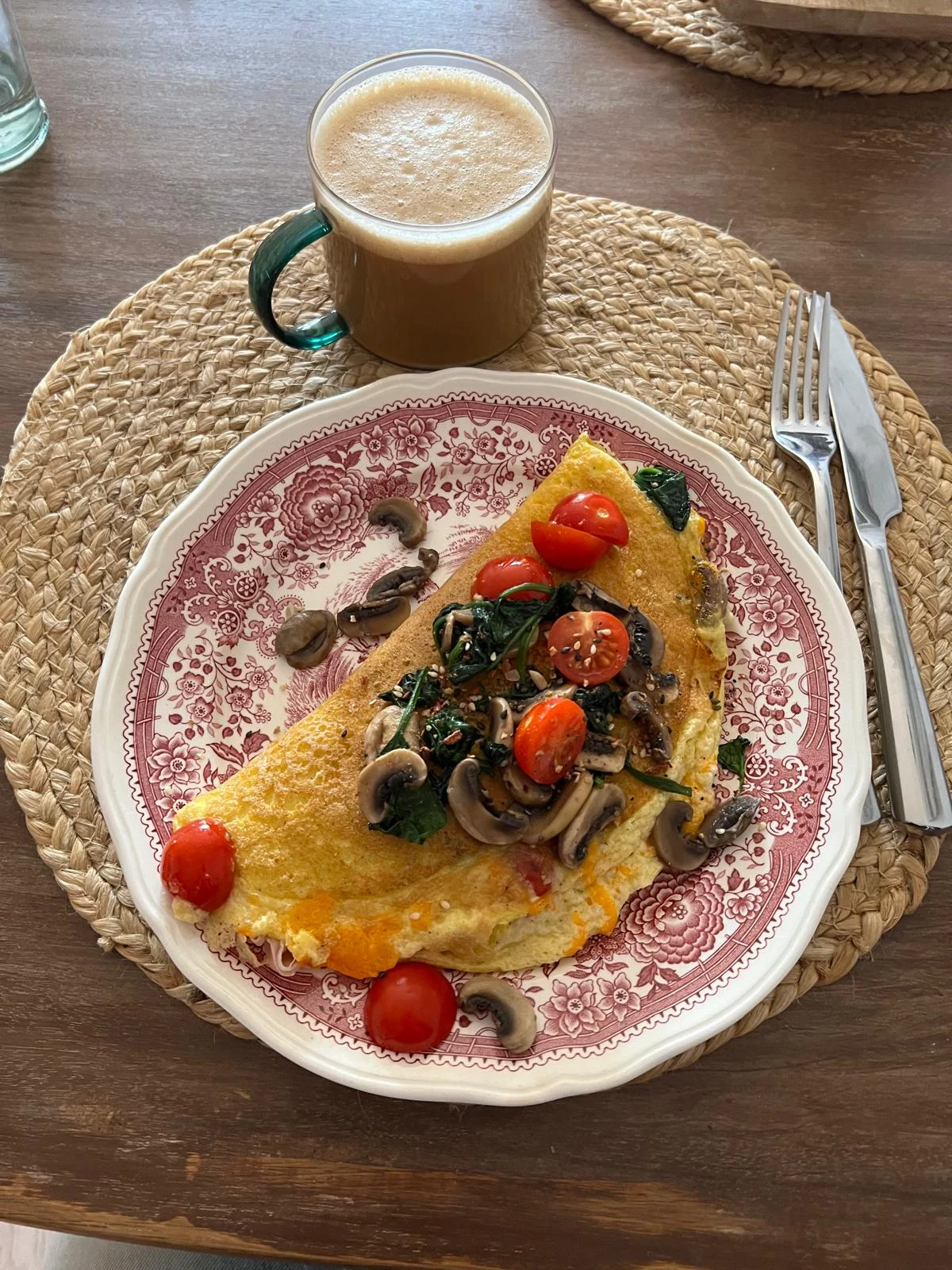 omlet z pomidorami koktajlowymi, szpinakiem i pieczarkami podany na ozdobnym talerzu, obok filiżanka kawy z mleczną pianką, sztućce ułożone po prawej stronie
