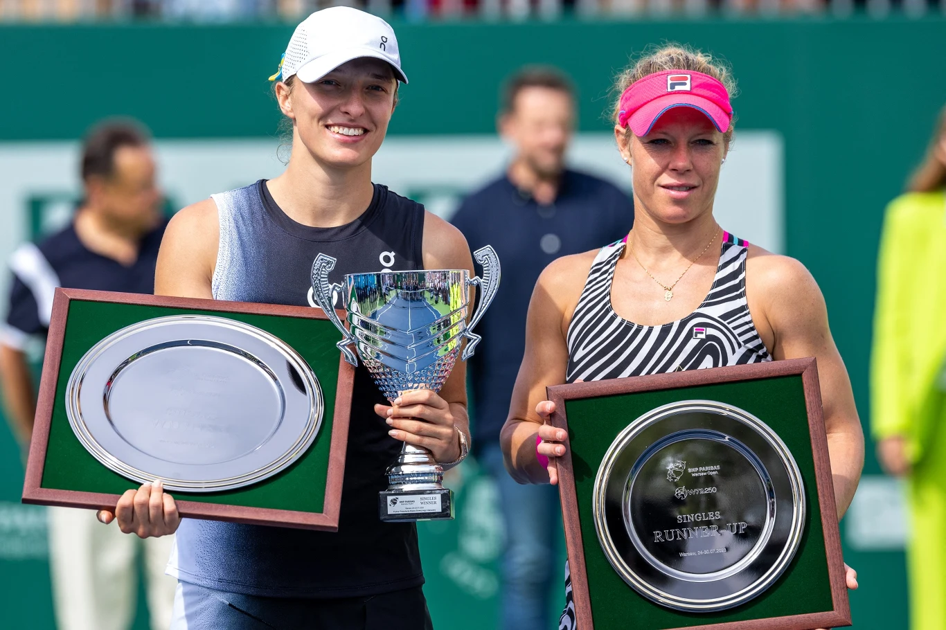 Iga Świątek i Laura Siegemund dwie tenisistki trzymają trofea i tablice pamiątkowe, jedna z nich uśmiechnięta z pucharem i srebrną tacą, druga z poważnym wyrazem twarzy prezentuje dużą plakietę, w tle rozmyte postacie oraz zielone ogrodzenie kortu tenisowego
