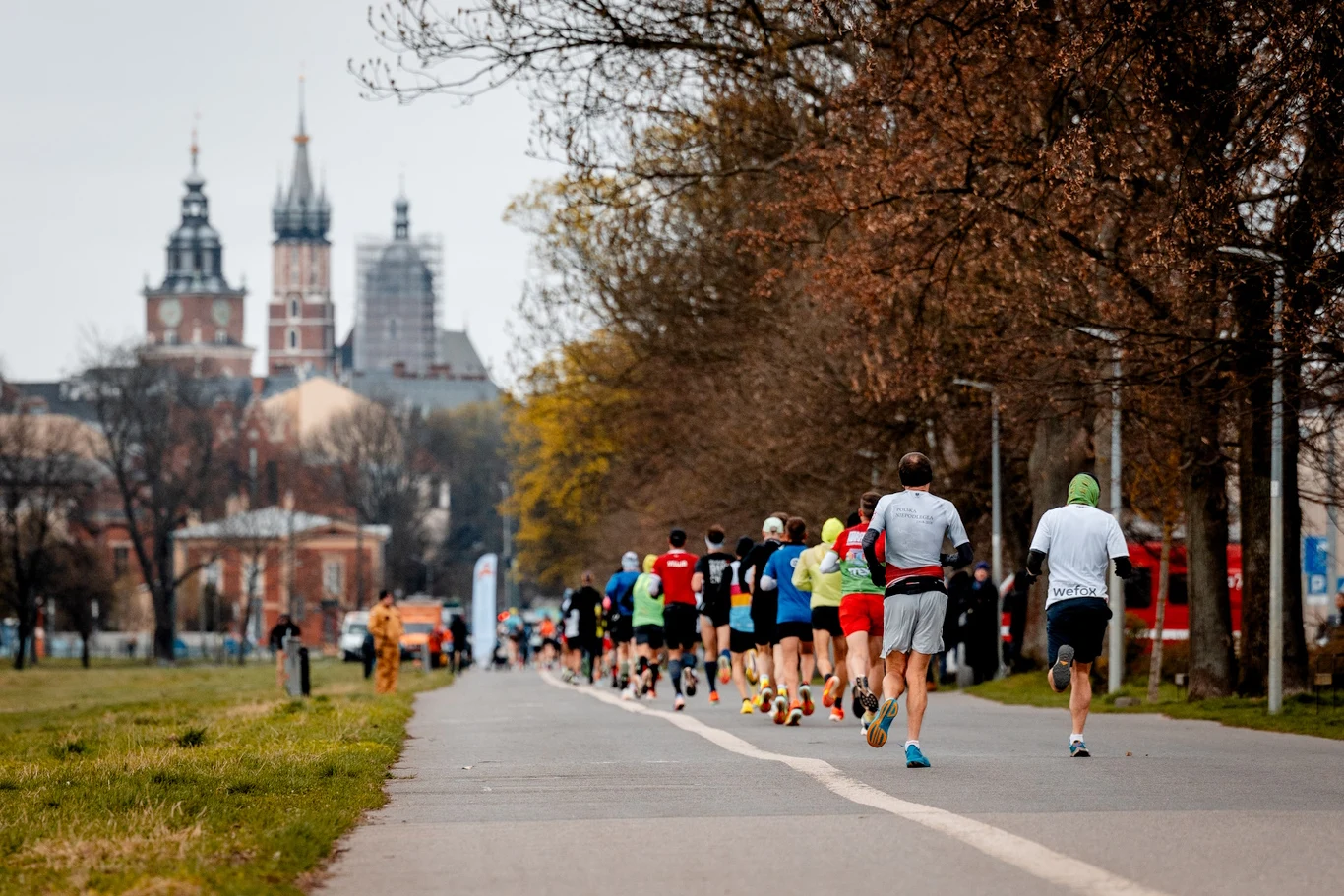 Grupa biegaczy uczestniczących w miejskim maratonie na asfaltowej alejce, otoczonej drzewami, w tle widoczna zabytkowa architektura z wysokimi wieżami kościoła.