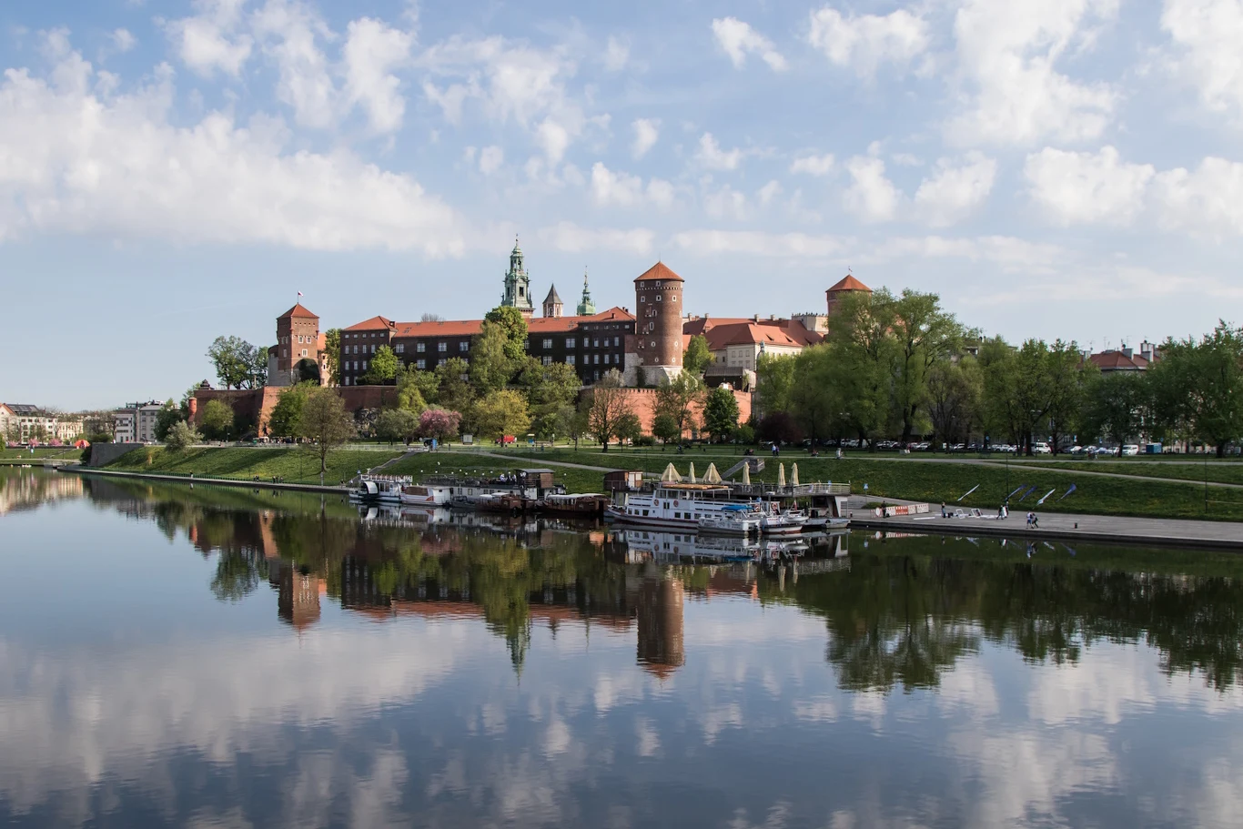 Zamek Wawel otoczony zielenią odbijający się w spokojnej tafli Wisły, zacumowane statki przy brzegu oraz spacerowicze na bulwarze.