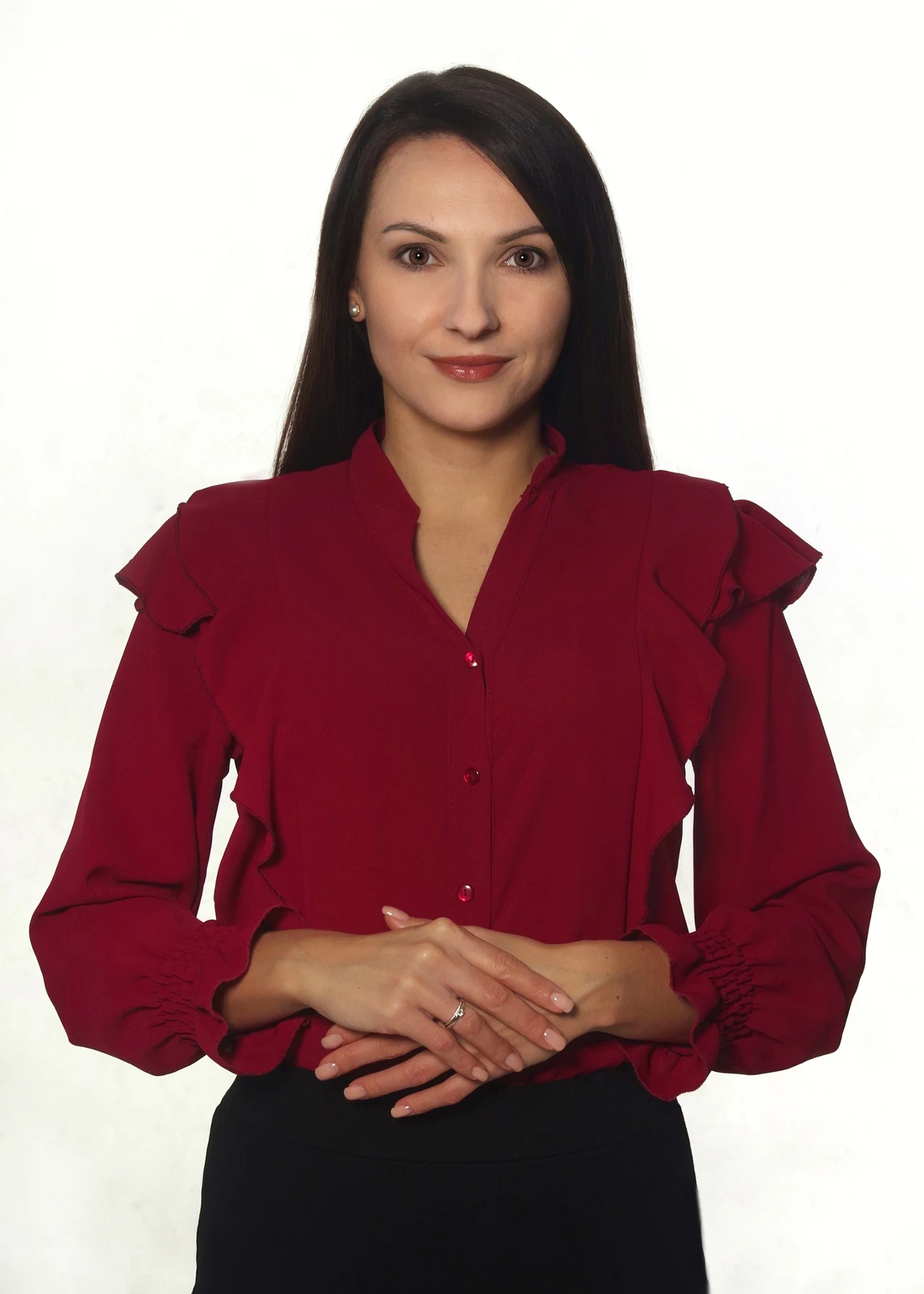 Kobieta o długich, prostych, ciemnych włosach w eleganckiej bordowej bluzce z falbanami, stojąca na jasnym tle z rękami ułożonymi jedna na drugiej na wysokości talii.