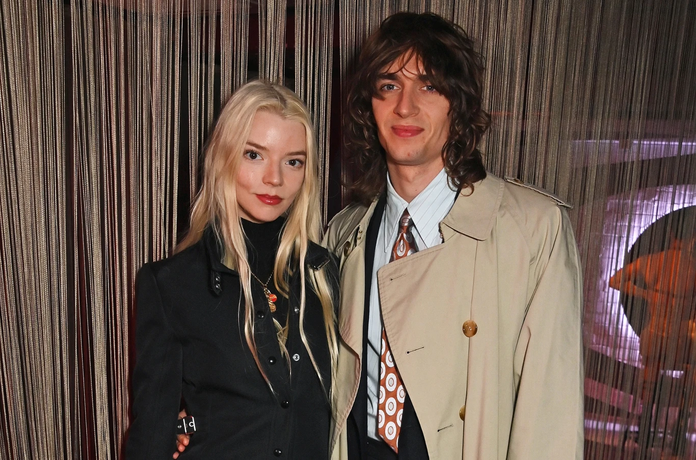 Anya Taylor Joy i Malcolm McRae Kobieta o długich blond włosach ubrana w czarny żakiet i mężczyzna z długimi brązowymi włosami w beżowym płaszczu oraz białej koszuli i wzorzystym krawacie stoją obok siebie na tle dekoracyjnych, pionowych sznurków.