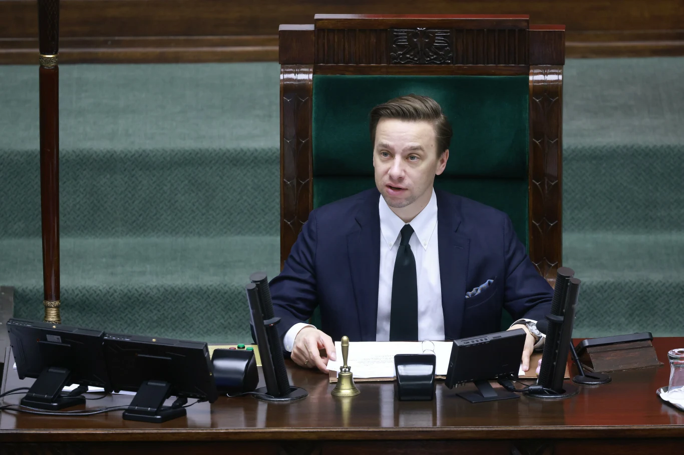 Wicemarszałek Sejmu Krzysztof Bosak Wicemarszałek Sejmu Krzysztof Bosak siedzi za zielonym biurkiem z mikrofonami i dokumentami, w tle sala obrad.