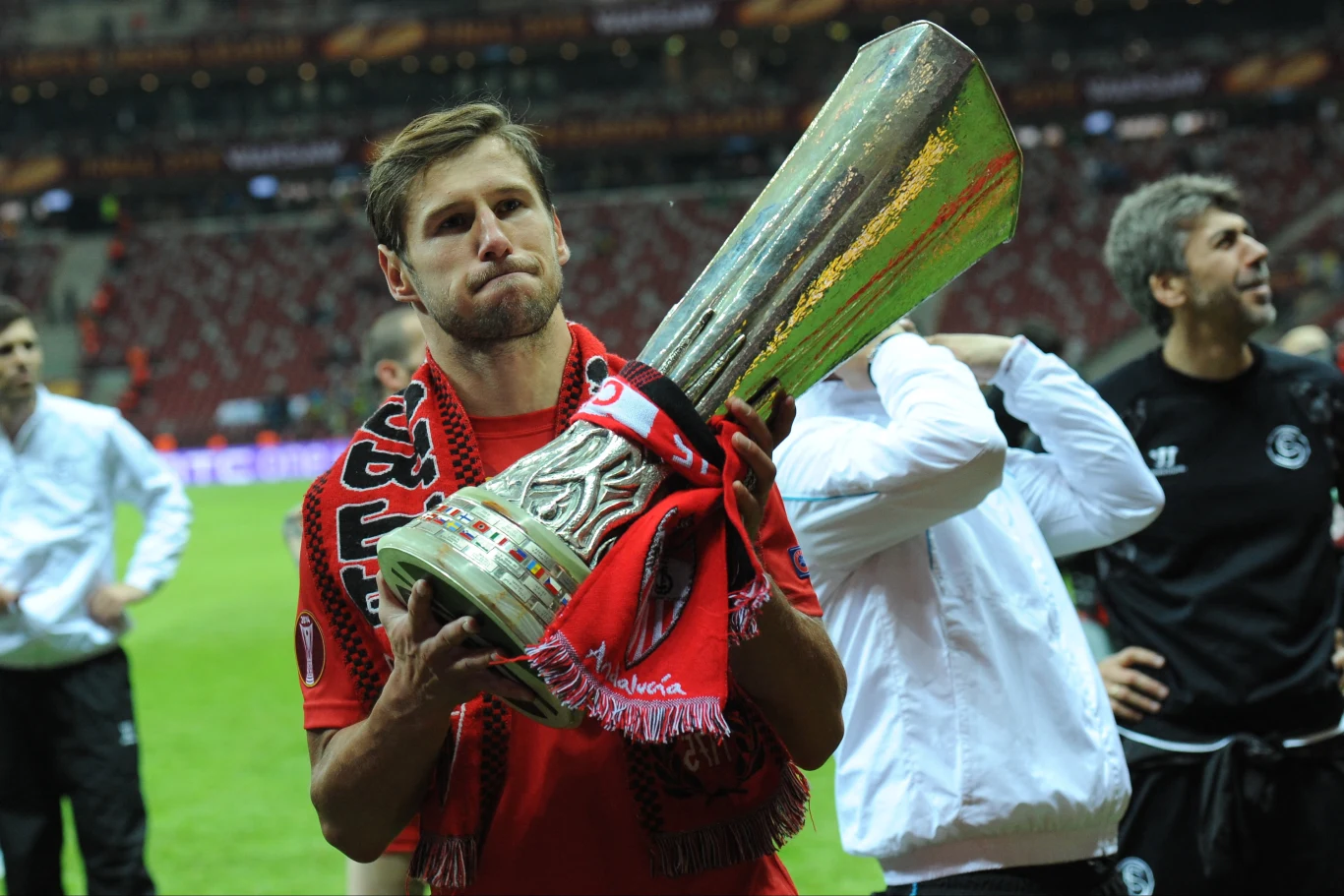 piłkarz w czerwonym stroju z szalikiem na szyi trzyma w dłoniach duży, srebrny puchar, wokół niego stoją inni zawodnicy na stadionie