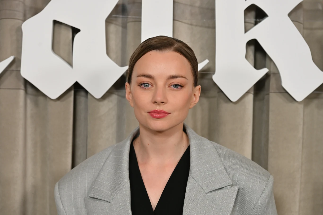 Kamila Urzędowska Kobieta z jasną cerą i jasnymi włosami spiętymi do tyłu, ubrana w jasnoszarą, oversize'ową marynarkę oraz czarny top, stoi na tle beżowych zasłon oraz dużych białych liter w stylu gotyckim.