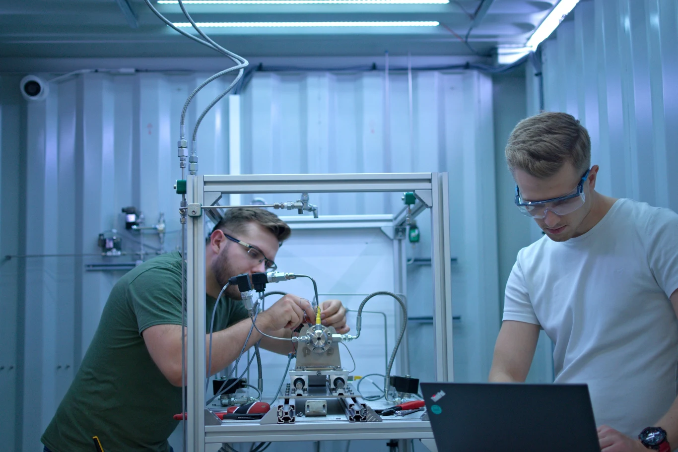 Dwóch mężczyzn w okularach ochronnych pracuje w laboratorium nad skomplikowanym urządzeniem mechanicznym; jeden skupia się na precyzyjnej regulacji maszyny, drugi analizuje dane na laptopie stojącym na metalowym stole otoczonym przewodami i narzędziami.