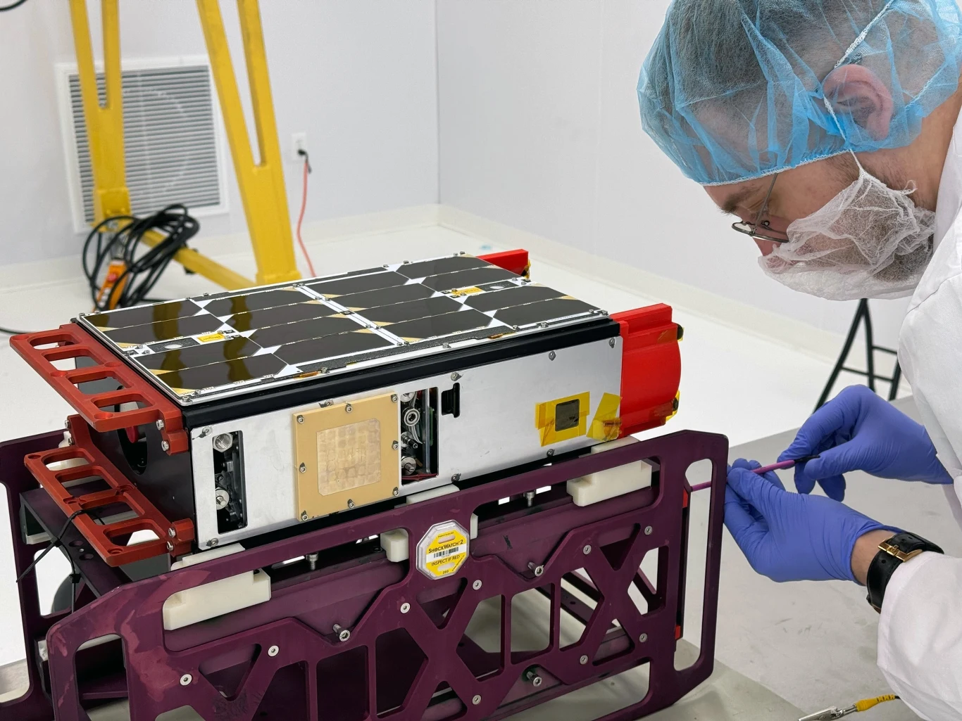 Inżynier w białym fartuchu, rękawiczkach i ochronnej siatce pracuje nad satelitą typu CubeSat umieszczonym na specjalnym stelażu w sterylnym laboratorium.
