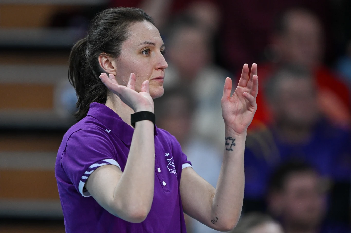 kobieta w fioletowej koszulce sportowej z uniesionymi dłońmi, skupiona podczas prowadzenia lub oceniania wydarzenia sportowego
