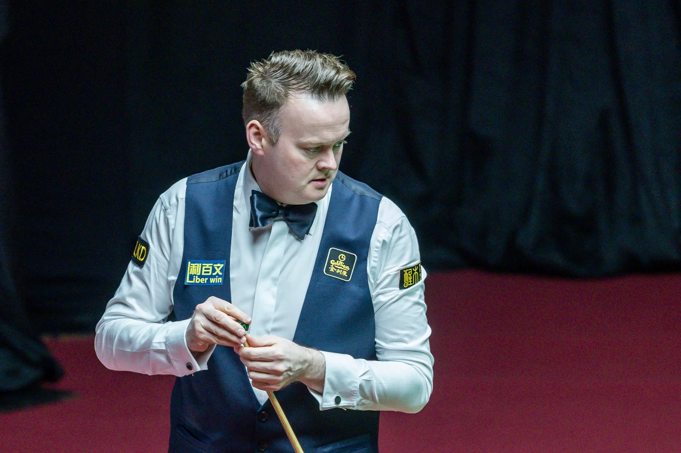 mężczyzna ubrany w elegancką kamizelkę i muchę trzyma kij bilardowy, stoi na tle czerwonego stołu snookerowego na profesjonalnym turnieju