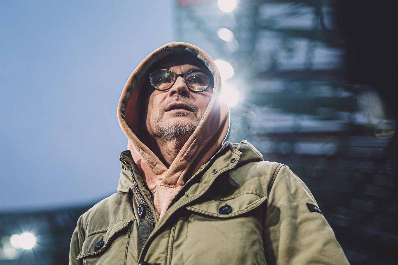 Wojciech Jagoda Mężczyzna w okularach i beżowej kurtce z kapturem, pod którym ma założoną bluzę z kapturem, stoi na tle jasnych świateł stadionu lub dużego obiektu, patrząc lekko w górę.