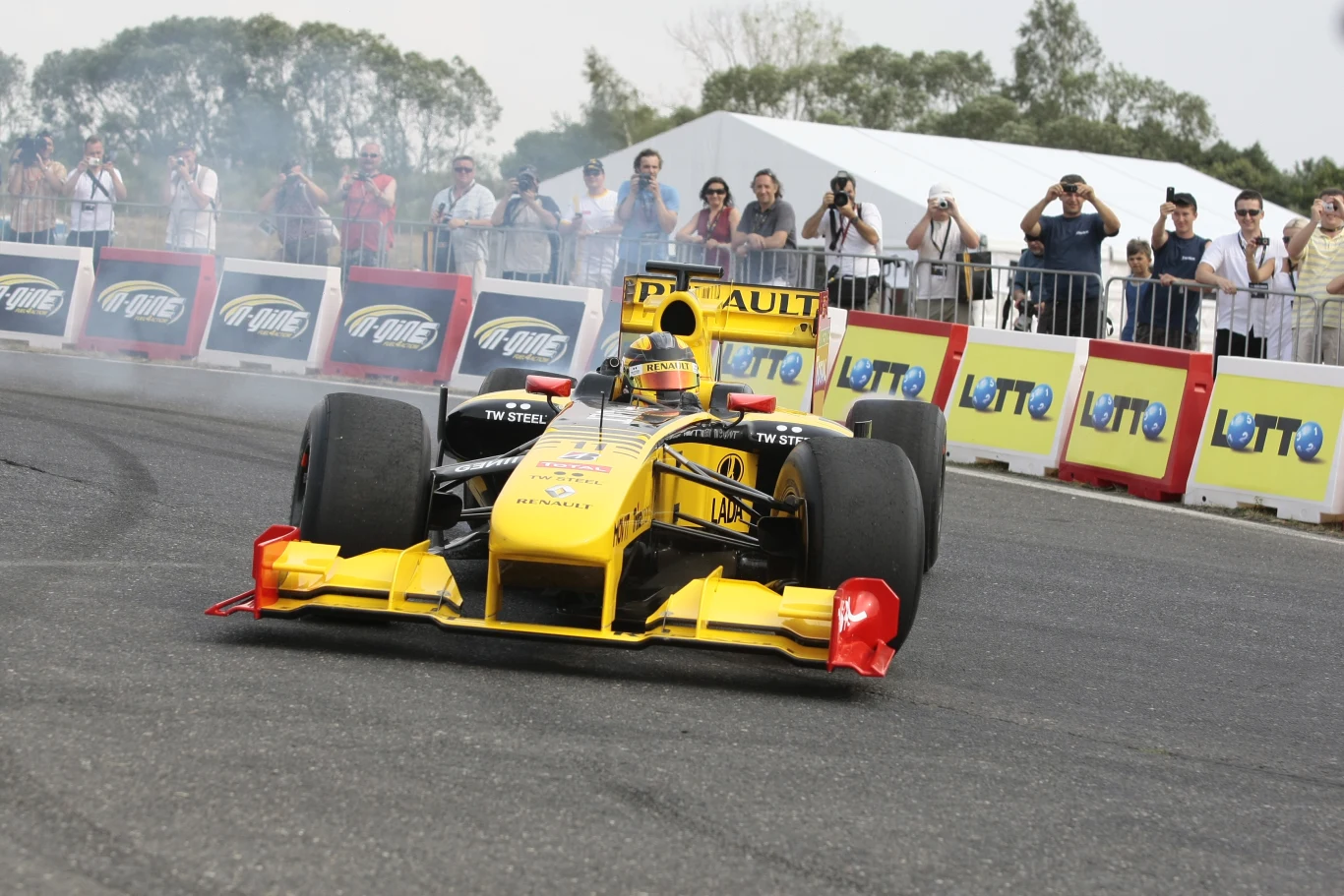 Robert Kubica w bolidzie F1 na Torze Poznań Wyścigowy bolid Renault w żółto-czarnych barwach wykonujący efektowny manewr na asfalcie, unoszący się dym spod opon i kibice fotografujący wydarzenie za barierkami z reklamami Lotto.