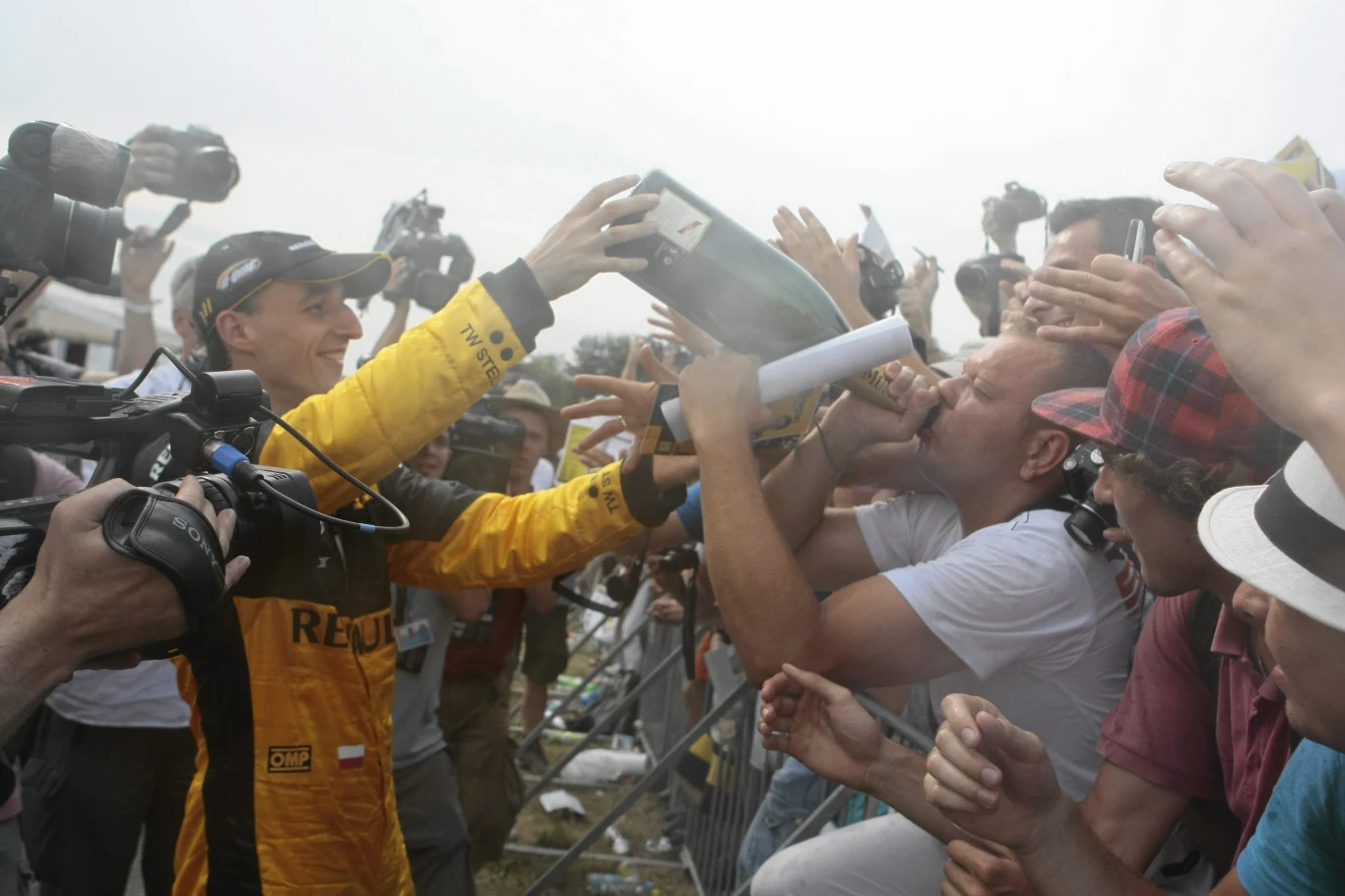 Robert Kubica świętuje z kibicami na Torze Poznań w 2010 roku Robert Kubica na Torze Poznań 2010, w żółtym kombinezonie Renault, podaje szampan kibicom przez barierkę.