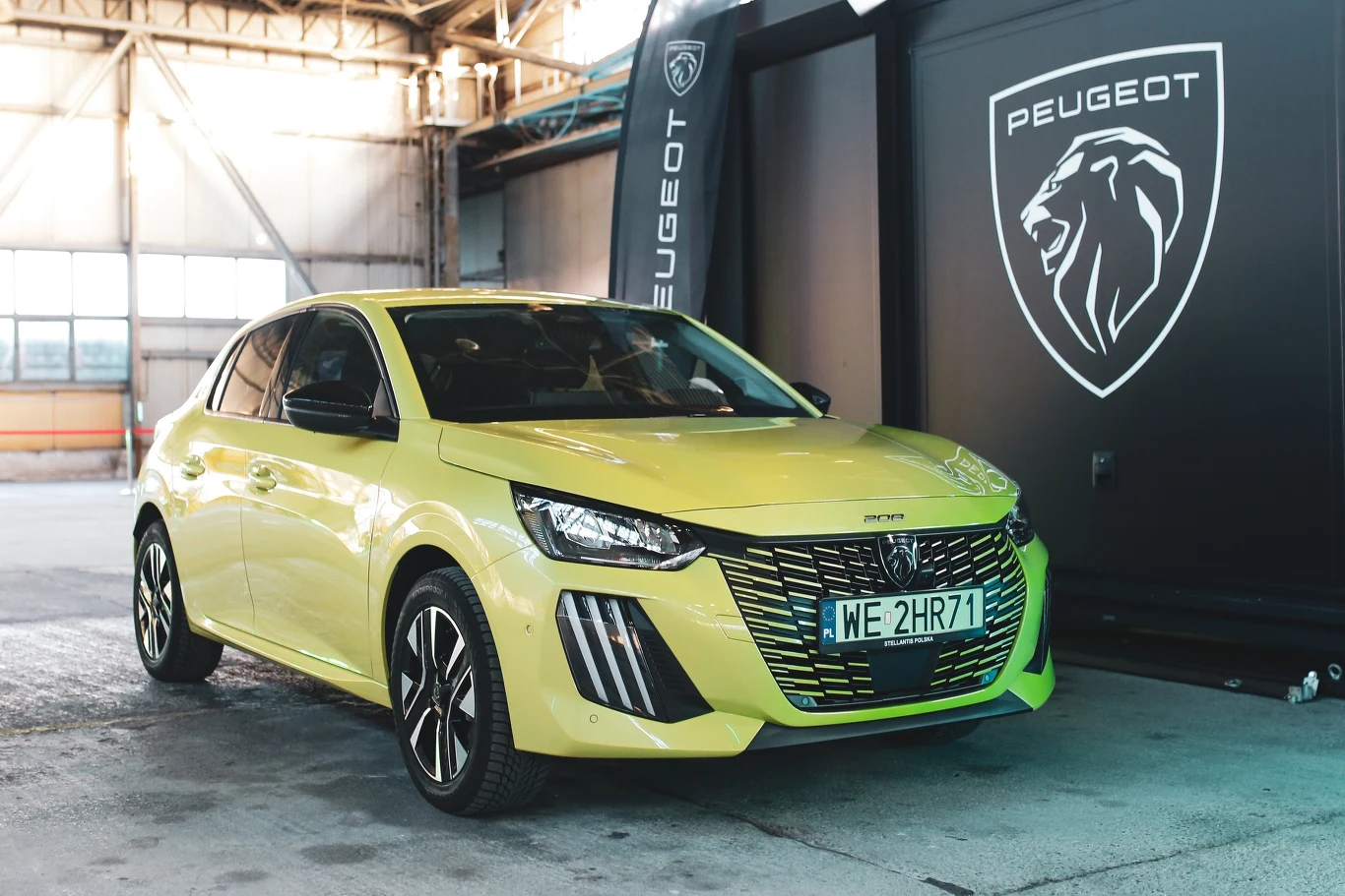 jasnożółty samochód Peugeot 208 ustawiony przodem w nowoczesnej hali, pod dużym banerem z logo marki i lwem
