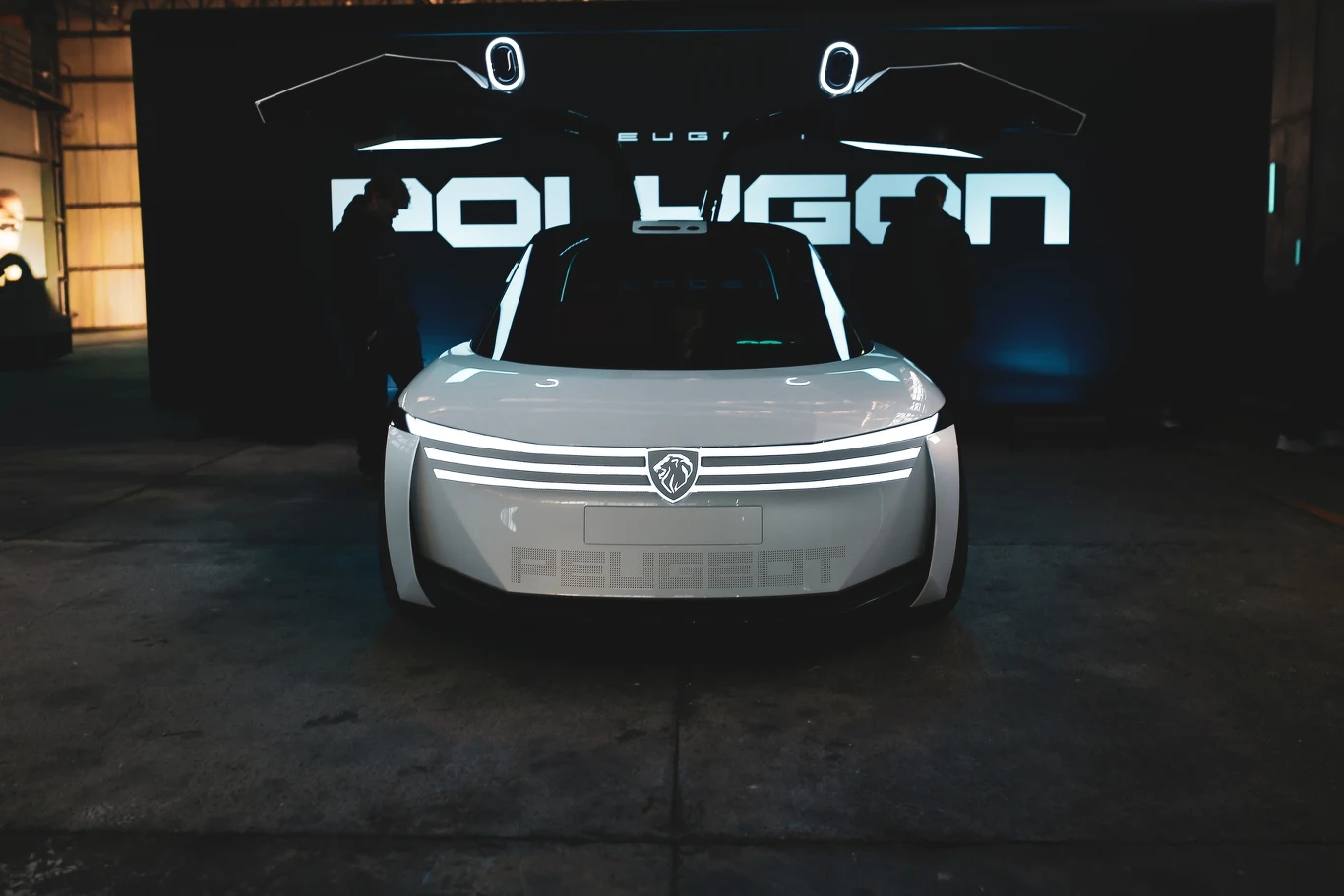 biały, futurystyczny samochód z otwartymi do góry drzwiami typu gullwing, mocno podświetlony front z poziomymi paskami LED, logo producenta na masce, ciemne otoczenie z widocznym podświetlonym napisem „POLYGON” w tle