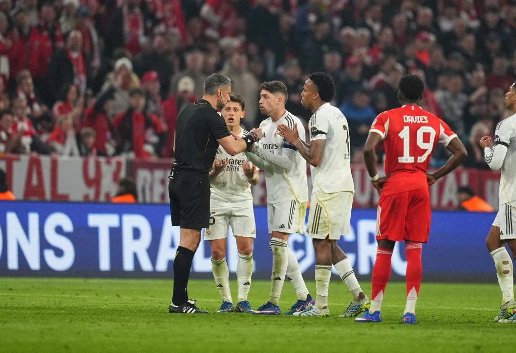 grupa piłkarzy w białych i czerwonych strojach dyskutuje z sędzią na murawie stadionu, wokół nich tłum kibiców w czerwonych barwach