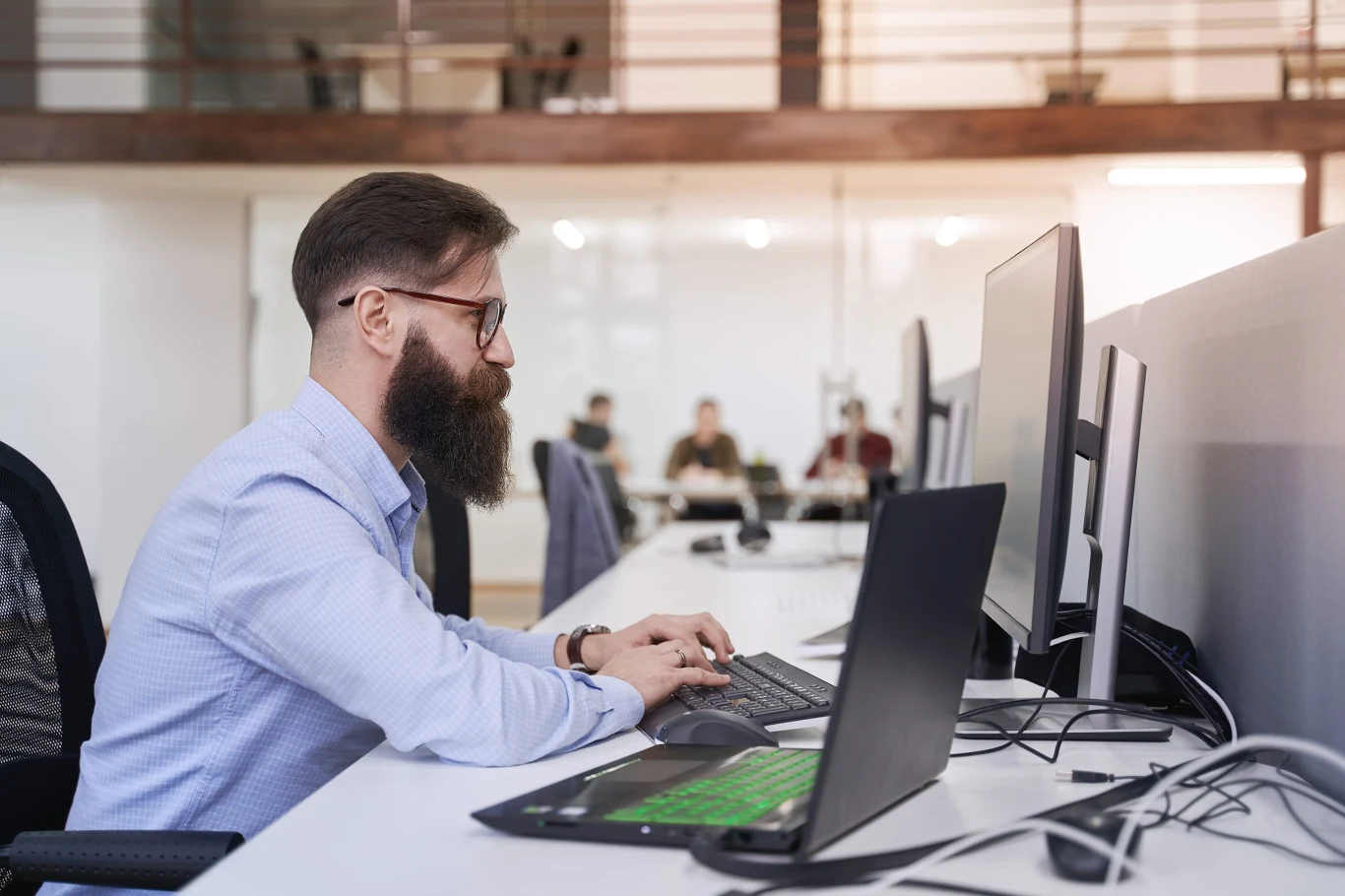 Model współpracy B2B od lat zyskuje na popularności, szczególnie w branżach wymagających wysokich kompetencji specjalistycznych. Mężczyzna z brodą i okularami pracujący przy monitorach i laptopie w nowoczesnym, otwartym biurze.