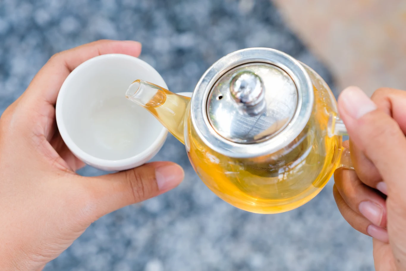 Herbata z suszonych liści popularnego drzewa przyspiesza metabolizm. Ręce nalewające herbatę z suszonych liści brzozy z przezroczystego dzbanka do białej filiżanki na marmurze.