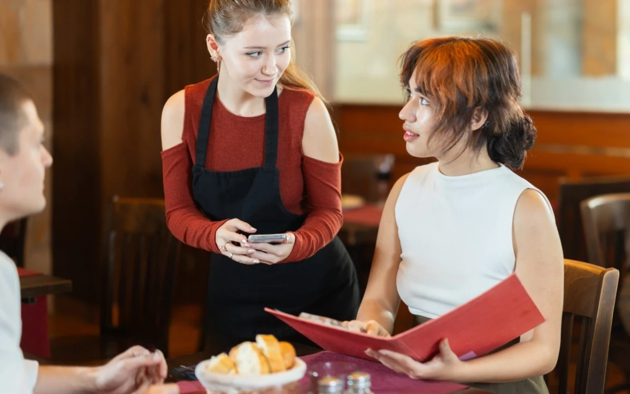 kelnerka uważnie przyjmuje zamówienie od kobiety siedzącej przy stole z otwartym menu, podczas gdy obok znajduje się mężczyzna z koszem pieczywa