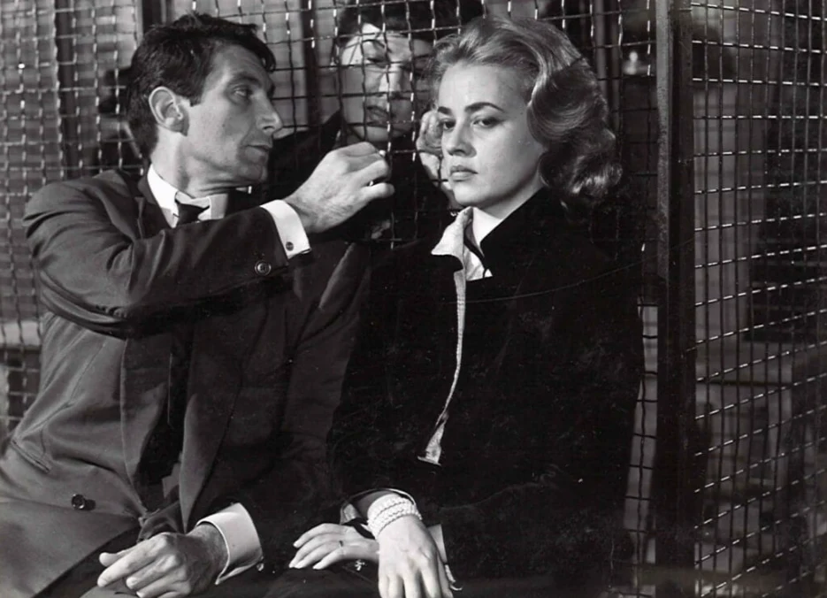 Kadr z filmu "Windą na szafot", fot. Timeless Film Festival Warsaw Mężczyzna w garniturze nachyla się w kierunku zamyślonej kobiety z blond włosami, siedzącej obok, za nimi widoczna siatka metalowa, co nadaje scenie napięcia i intymności.