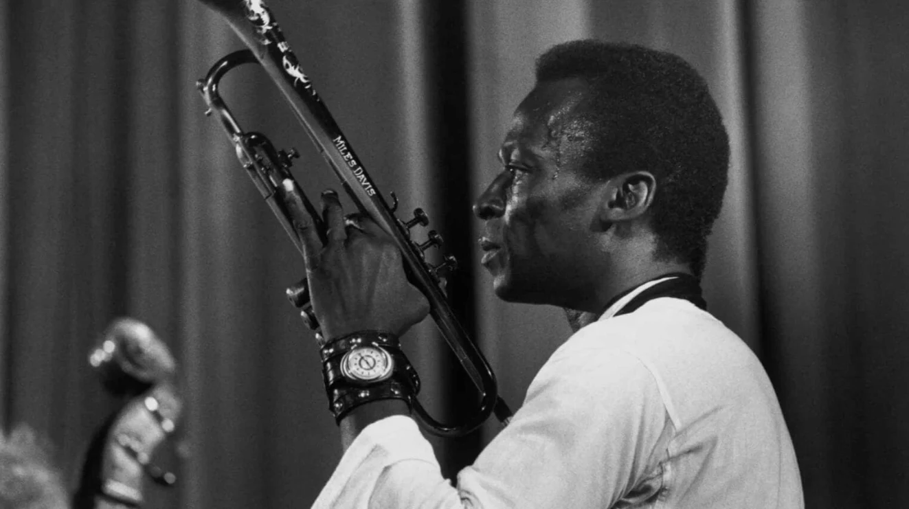 Kadr z dokumentu "Miles Davis: Birth of the Cool", fot. Timeless Film Festival Warsaw Muzyk trzymający trąbkę w dłoniach, ubrany w jasną koszulę i ozdobiony zegarkami na nadgarstkach, na tle zasłon scenicznych.