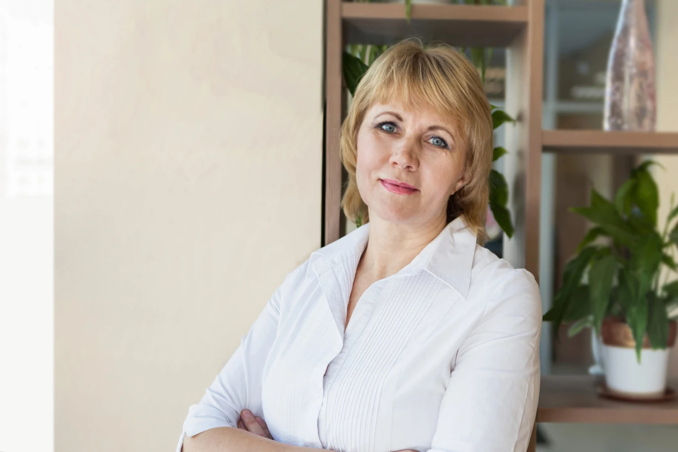 Kobieta w średnim wieku z blond włosami w białej koszuli stoi z założonymi rękami na tle półek z zielonymi roślinami doniczkowymi, patrzy spokojnie w stronę aparatu.