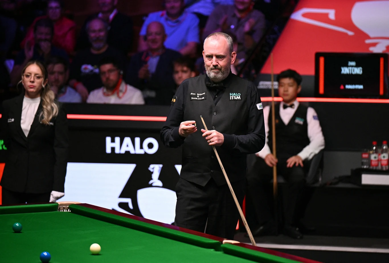 Mark Wiliams w finale MŚ w 2025 roku mężczyzna w czarnej kamizelce trzyma kij bilardowy, stojąc przy stole do snookera ze skupionym wyrazem twarzy; obok stoi sędzina w białej koszuli i czarnej marynarce, w tle publiczność oraz drugi zawodnik przygotowujący się do gry