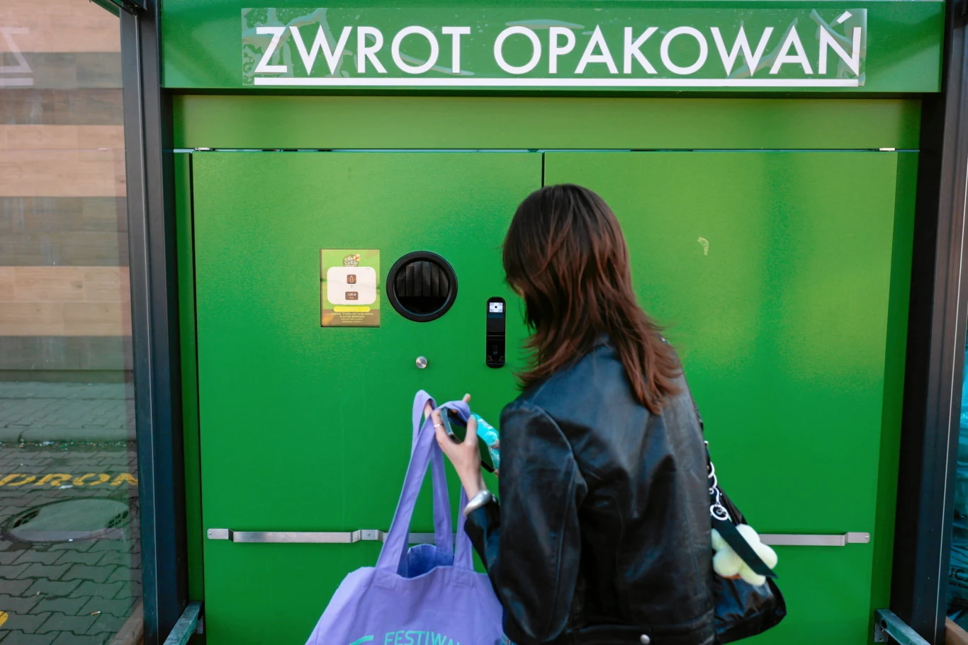 System kaucyjny budzi wiele emocji. Niektórzy widzą w zwrocie opakowań coś niegodnego ich poziomu Kobieta w skórzanej kurtce przy zielonym automacie do zwrotu opakowań z torbą butelek w ręku