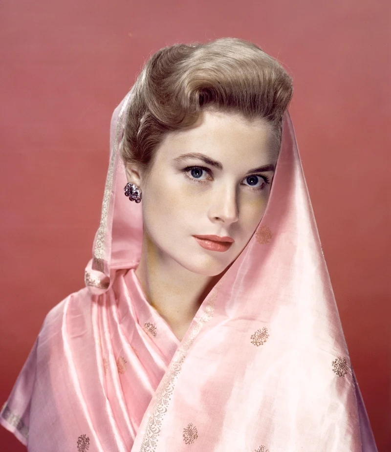 Kobieta o jasnej cerze i blond włosach spiętych w elegancką fryzurę, ubrana w jedwabny, różowy szal ozdobiony delikatnym wzorem, patrząca wprost z subtelnym uśmiechem na twarzy, tło monochromatyczne w odcieniu różu.