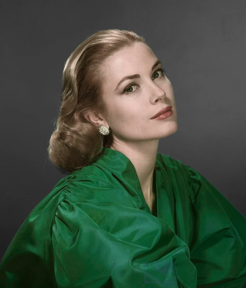 Elegancka kobieta o jasnej karnacji i blond włosach upiętych w stylowe fale, ubrana w zieloną, satynową bluzkę z bufiastymi rękawami, patrząca spokojnie w stronę obiektywu; całość dopełniają delikatny makijaż oraz efektowne kolczyki typu klipsy.