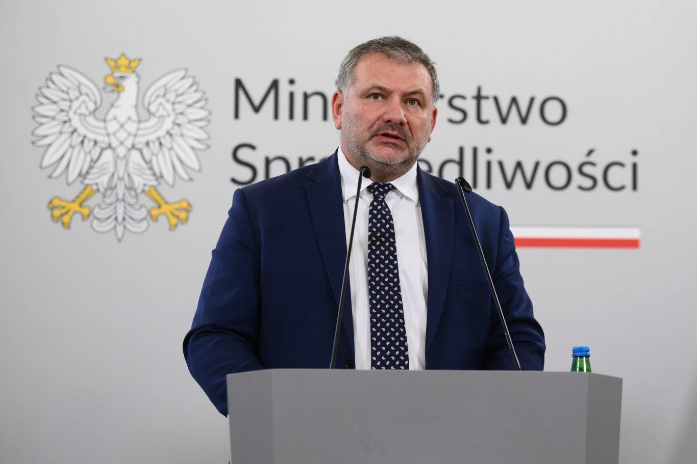 Minister sprawiedliwości Waldemar Żurek Waldemar Żurek przy mównicy, w tle herb Polski i napis 'Ministerstwo Sprawiedliwości', obok butelka wody.