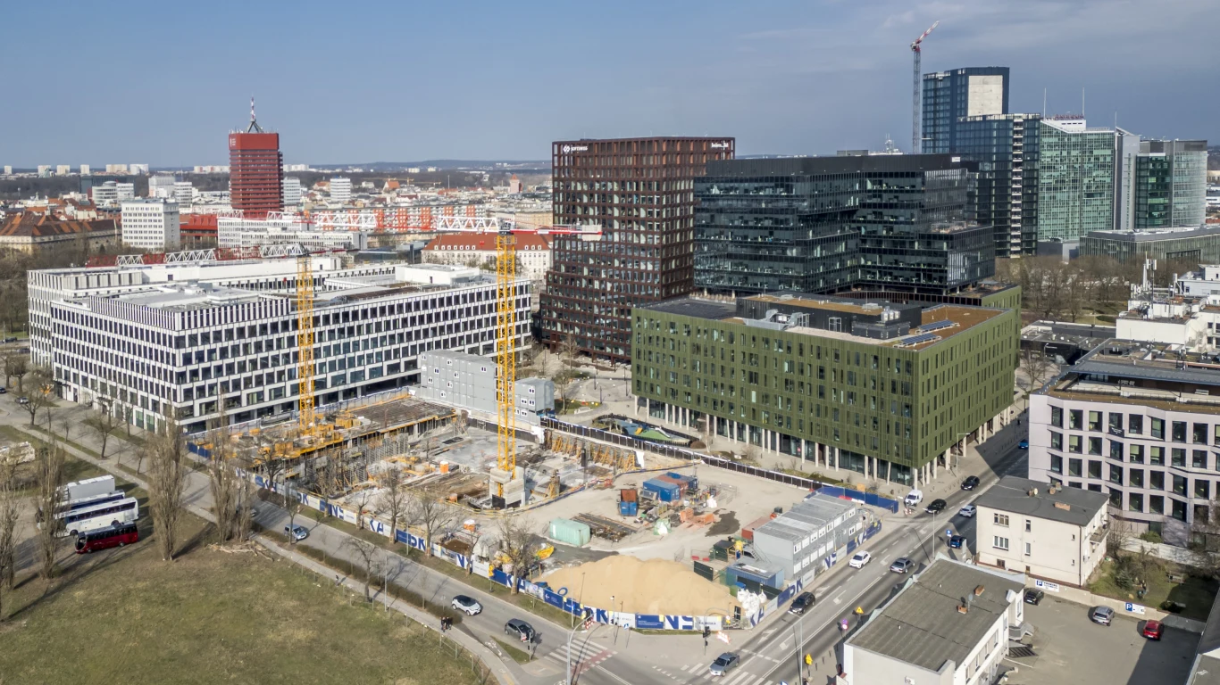 Certyfikat dla powstającego budynku został wydany na podstawie opinii technicznej wykonanej przez grupę specjalistów z Centrum Budownictwa Ochronnego, funkcjonującego w ramach Wydziału Inżynierii Lądowej i Geodezji Wojskowej Akademii Technicznej. nowoczesny kompleks biurowców z wyraźnie widoczną budową na pierwszym planie, gdzie pracują dwa żółte żurawie wieżowe; otoczenie tworzą wysokie budynki biurowe w różnych kolorach, a w tle rozciąga się panorama miasta