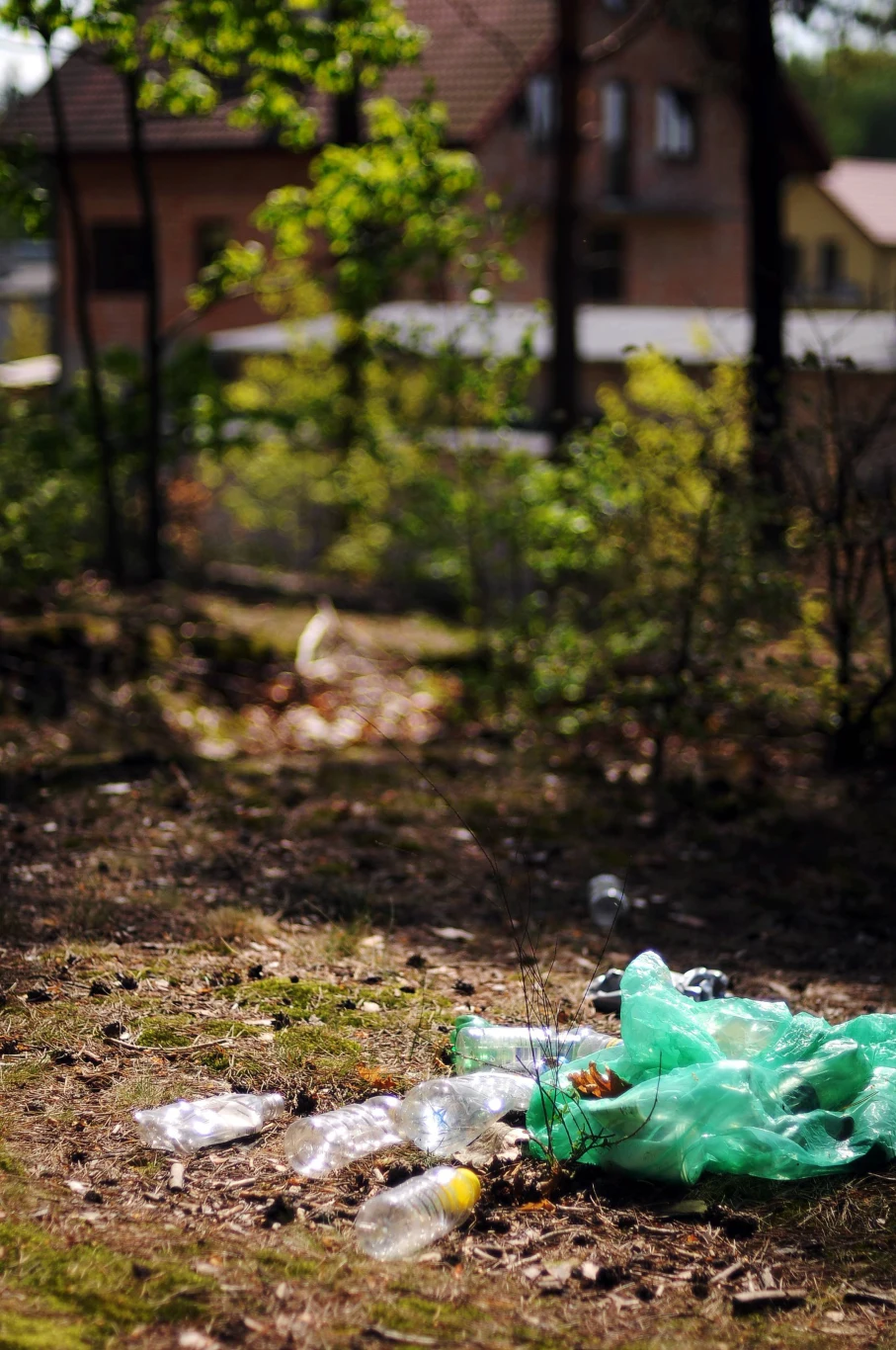 porzucone plastikowe butelki i zielony worek leżące na ziemi w lesie, w tle widoczne są drzewa oraz budynki mieszkalne