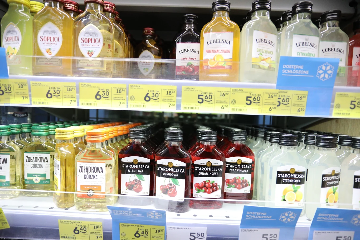 kolorowe butelki alkoholi różnych marek ustawione na półkach sklepowych, pośród nich widoczne etykiety Soplica, Lubelska i Staropolska, pod butelkami żółte i niebieskie etykiety cenowe
