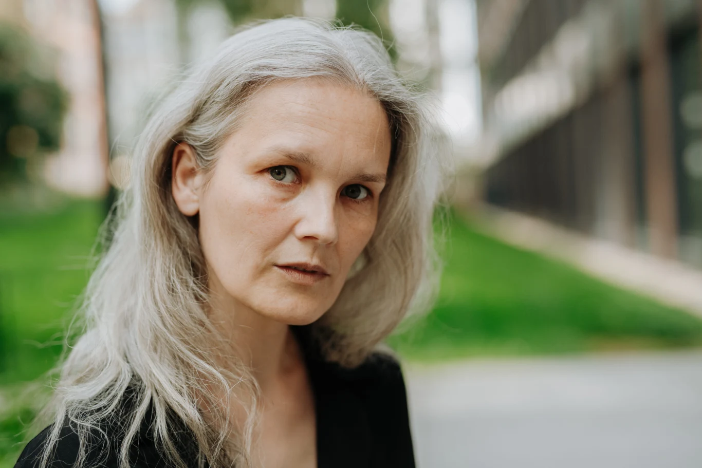 Kto najczęściej wywołuje burzę w relacjach? Zodiakalni miłośnicy dramatu Kobieta o siwych, długich włosach i jasnej cerze patrzy poważnie w stronę obiektywu; w tle rozmyte zielone i miejskie elementy krajobrazu.