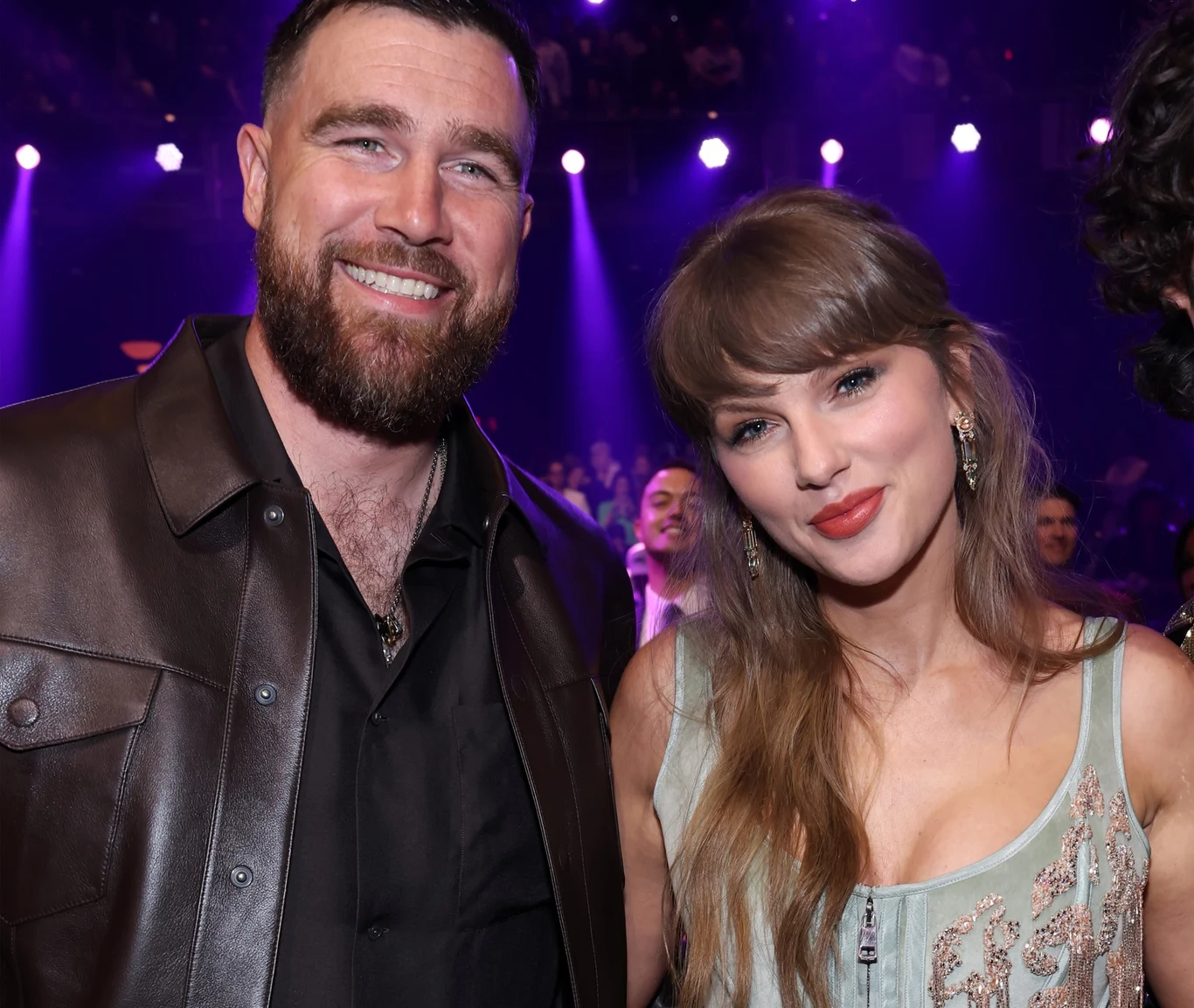 Taylor Swift i Travis Kelce biorą ślub. Przygotowania idą pełną parą Taylor Swift w jasnej sukni obok Travisa Kelce w skórzanej kurtce, na tle sceny i tłumu pod fioletowym światłem.