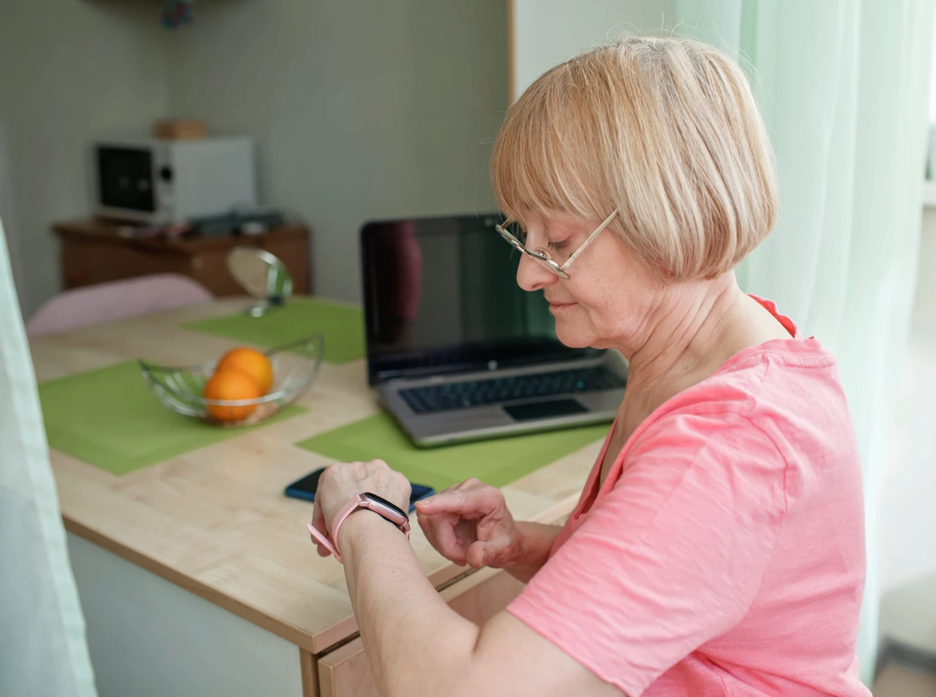 Pechowe i szczęśliwe dni w tygodniu. Kiedy warto działać, a kiedy uważać? starsza kobieta w okularach siedzi przy stole, sprawdzając opaskę elektroniczną na nadgarstku; na stole znajduje się laptop oraz miska z pomarańczami