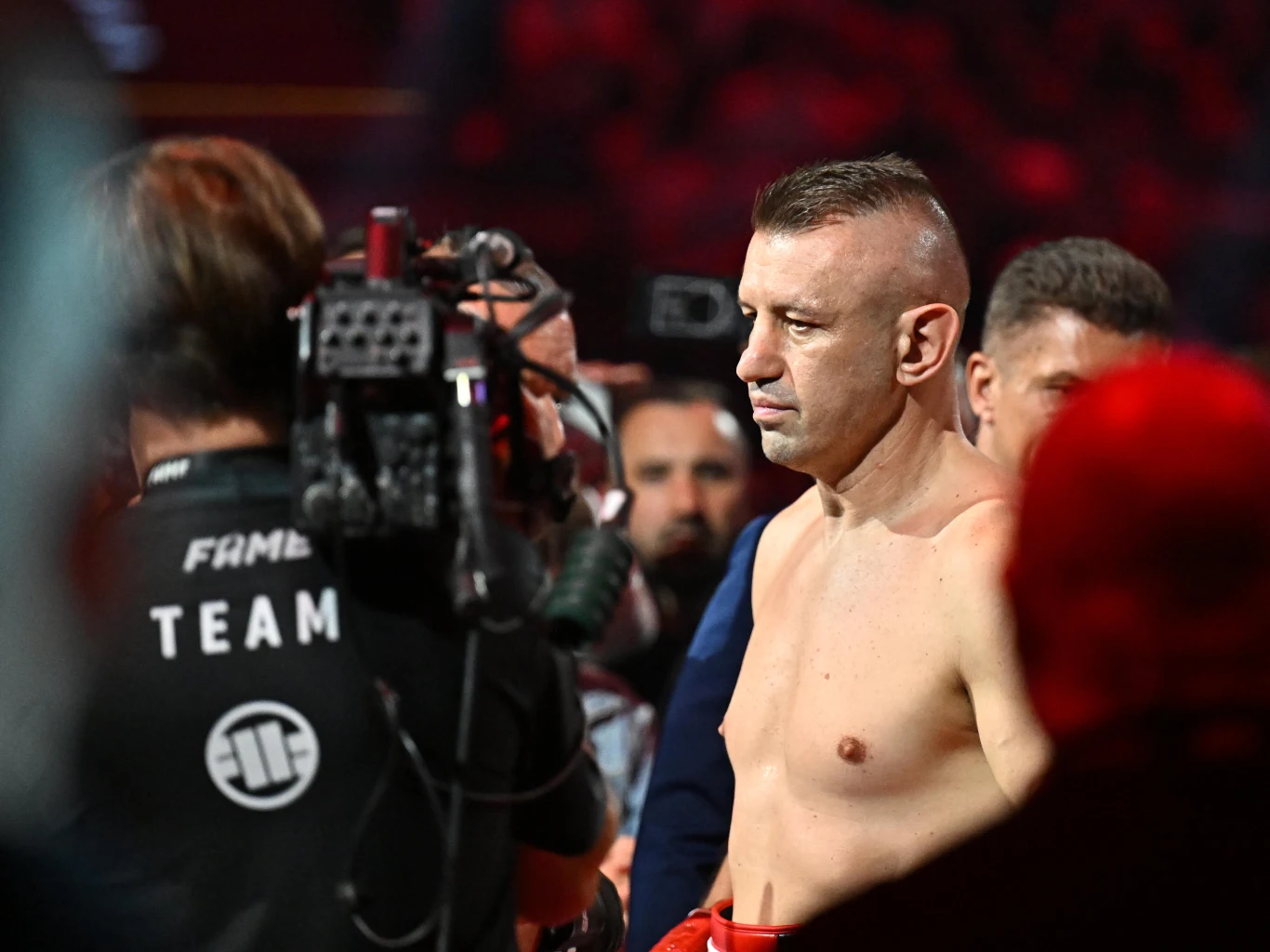Tomasz Adamek Skupiony zawodnik w rękawicach bokserskich stoi w jasnym oświetleniu ringu, otoczony przez członków ekipy i operatora kamery w czarnym stroju z napisem 'TEAM'.