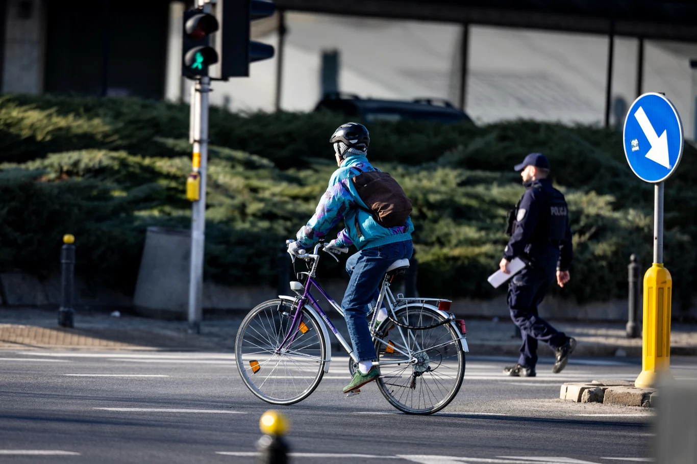 Jazda rowerem podlega tym samym regulacjom, co poruszanie się innymi pojazdami po drodze. rowerzysta w kolorowej kurtce i kasku przejeżdża przez skrzyżowanie na zielonym świetle, obok policjant, w tle znak i zieleń
